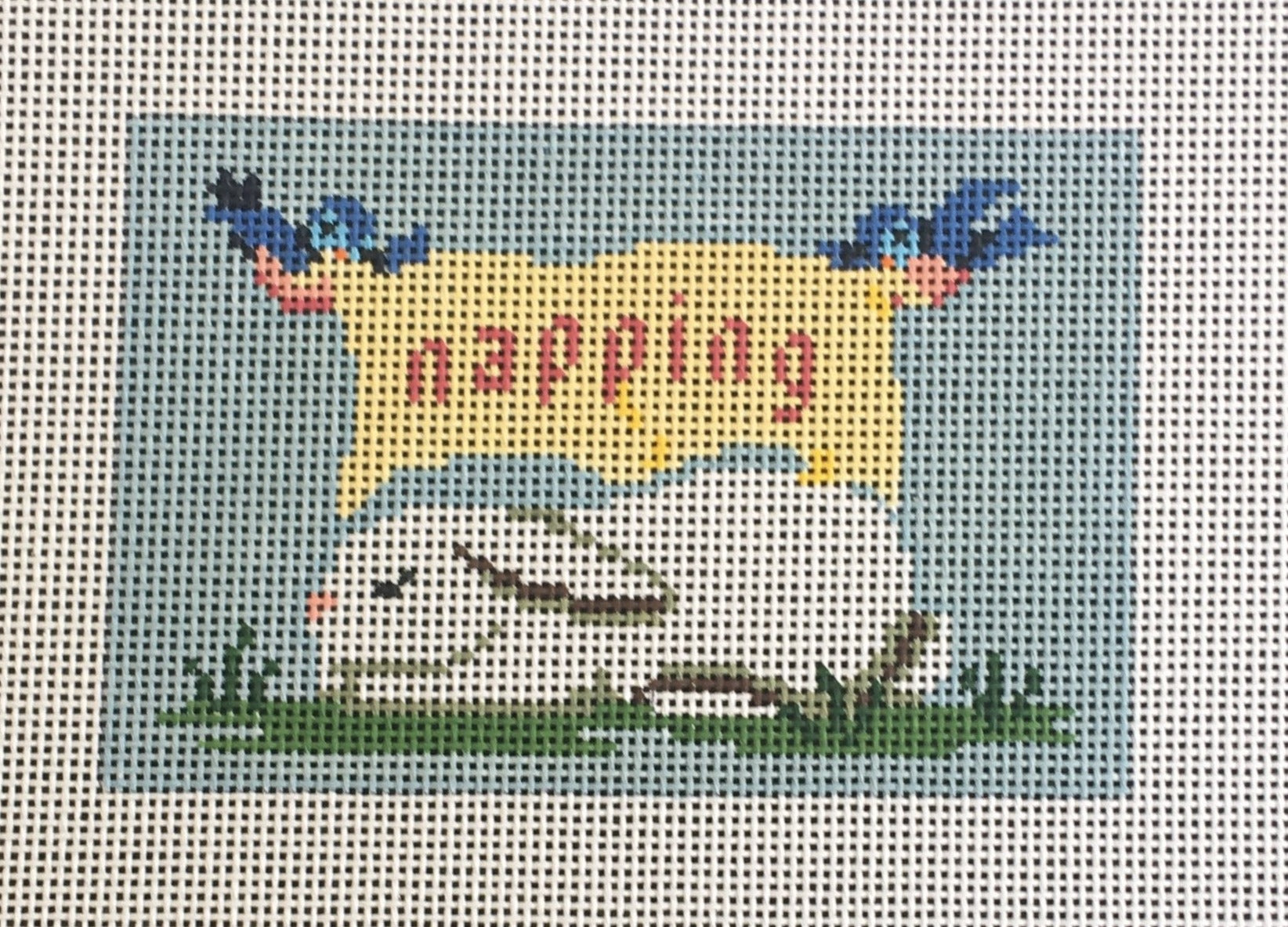 Susan Roberts needlepoint canvas sign for a child sleeping. Shows a sleeping bunny laying in the grass with two birds flying above holding a banner that says "napping"
