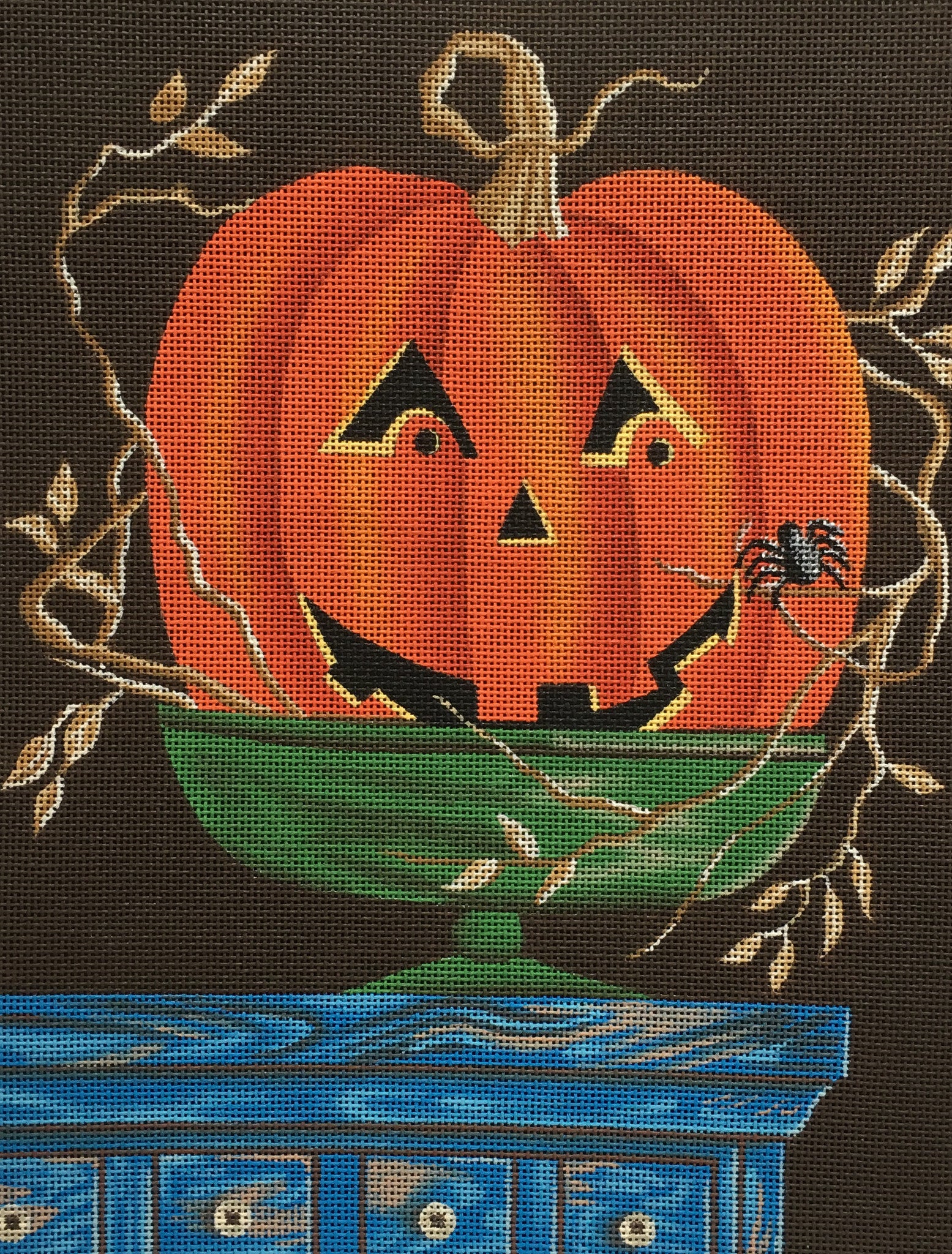 Kim Leo needlepoint canvas of a smiling jack-o-lantern in a green bowl on a farmhouse chest with a small spider