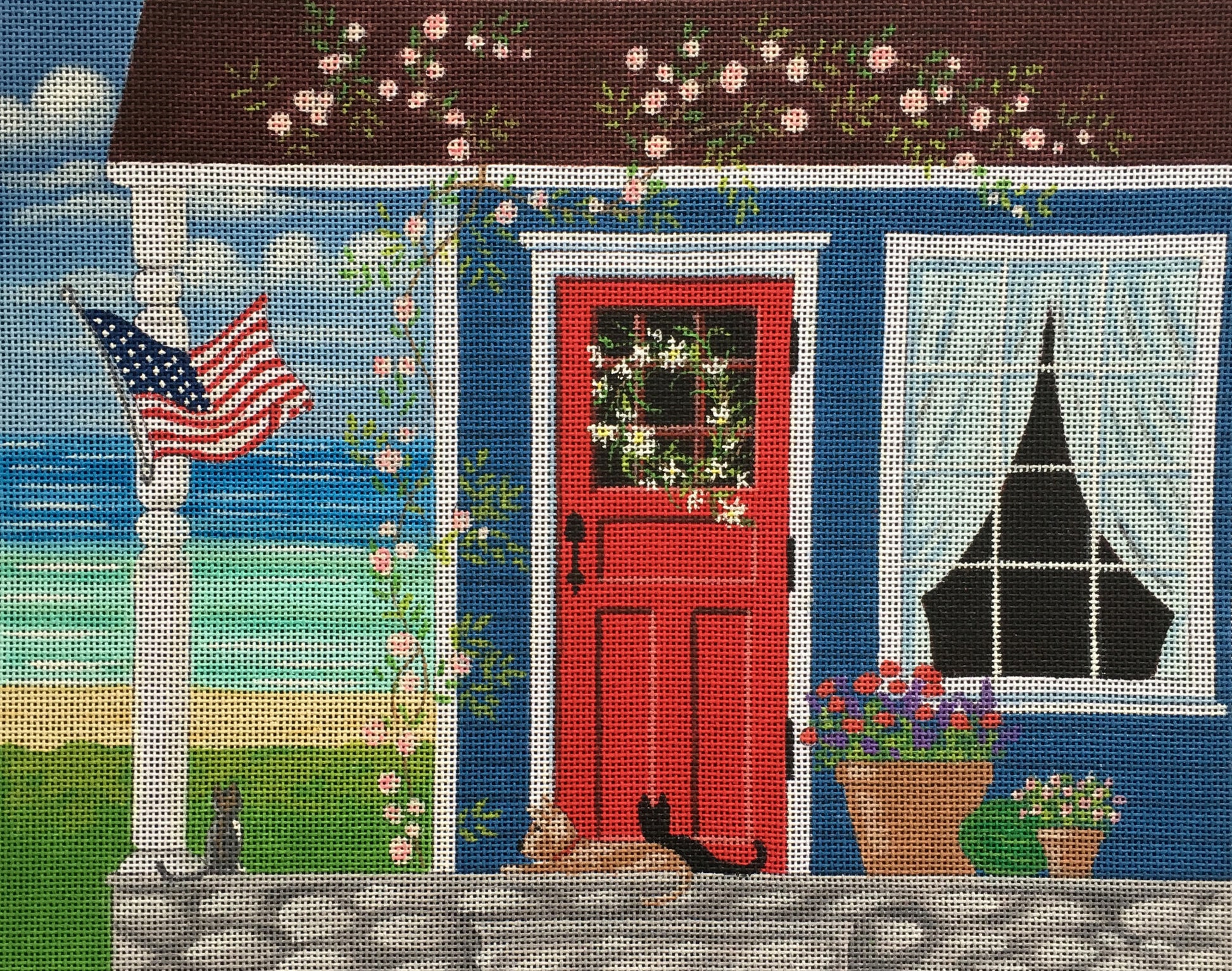 Kim Leo needlepoint canvas of a blue house with a porch and an American flag by the sea with cats and a dog